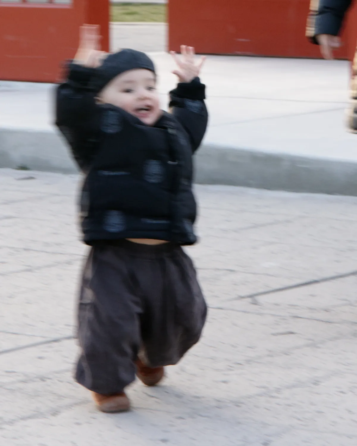 A toddler running with raised hands appears blurred by motion in front of red doors.