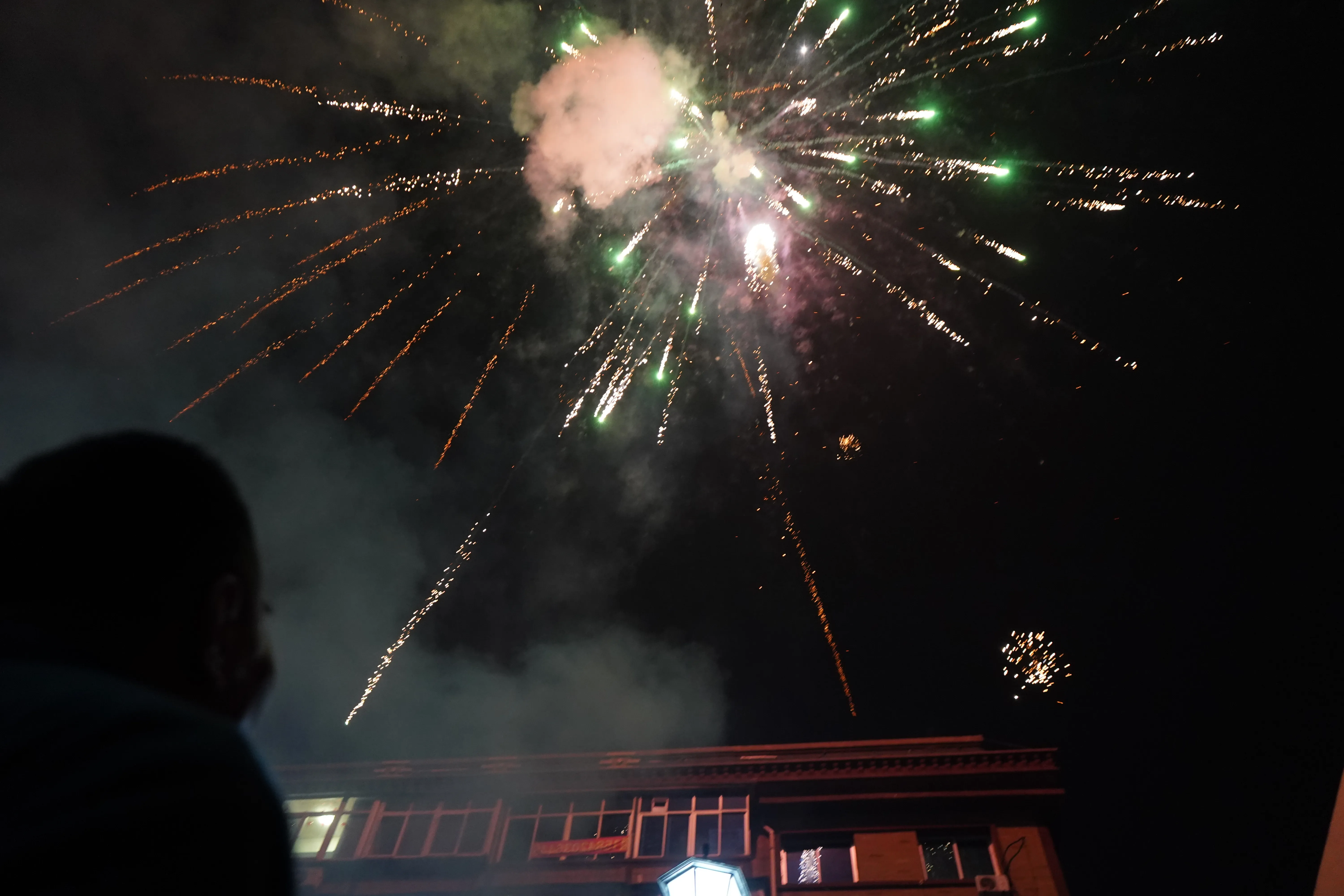 Silhouetted figure watching fireworks overhead as smoke drifts across the courtyard between apartment buildings.
