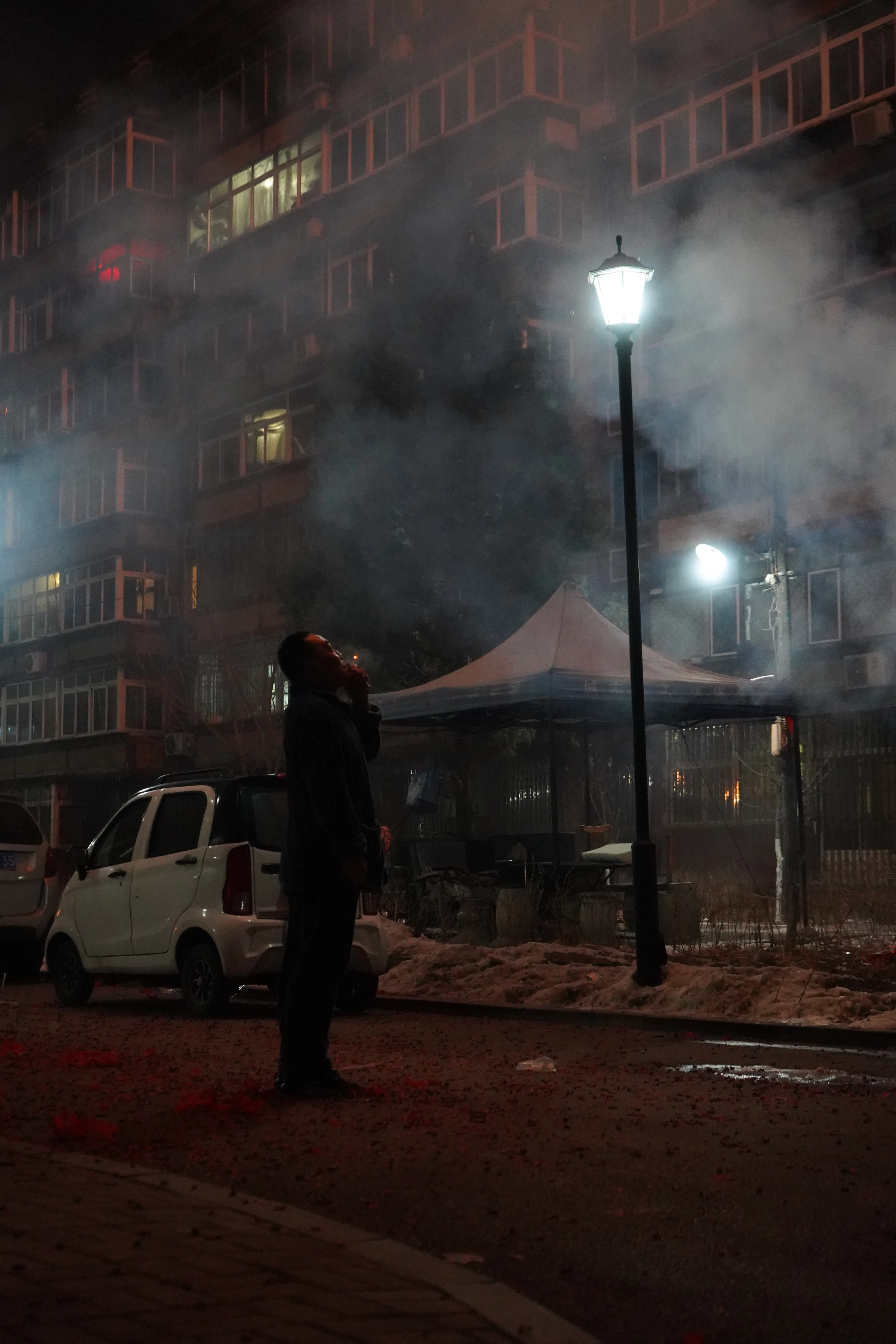 Man taking a drag on a cigarette while gazing at fireworks lighting up the smoky night sky.
