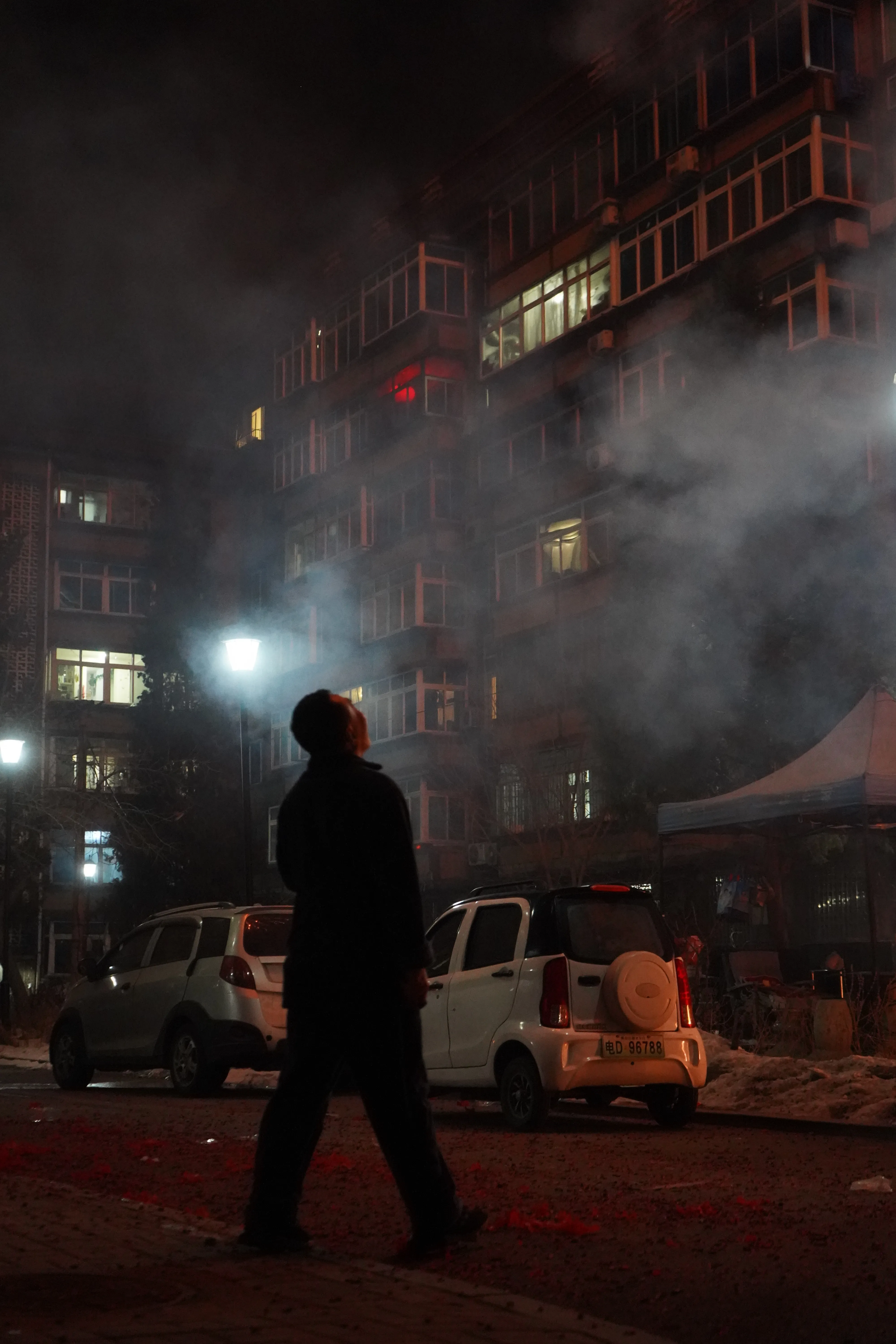 Man standing in a residential courtyard looking up at fireworks exploding above the buildings.